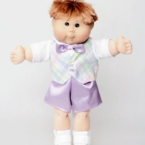 May include: A Cabbage Patch Kid doll dressed in a formal outfit. The doll has auburn hair and is wearing a white shirt, a plaid vest with pastel colors, a purple bow tie, and matching purple satin shorts. White sneakers complete the look.