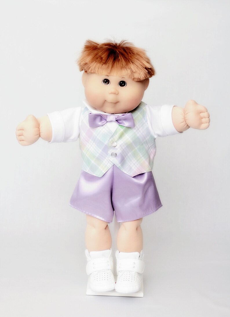 May include: A Cabbage Patch Kid doll dressed in a formal outfit. The doll has auburn hair and is wearing a white shirt, a plaid vest with pastel colors, a purple bow tie, and matching purple satin shorts. White sneakers complete the look.