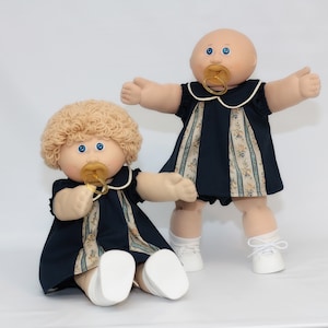 May include: Two Cabbage Patch Kids dolls. One doll is seated, the other is standing. Both dolls have blue eyes, pacifiers, and wear navy blue dresses with floral accents. The seated doll has blonde curly hair, while the standing doll is bald. Both dolls wear white shoes.