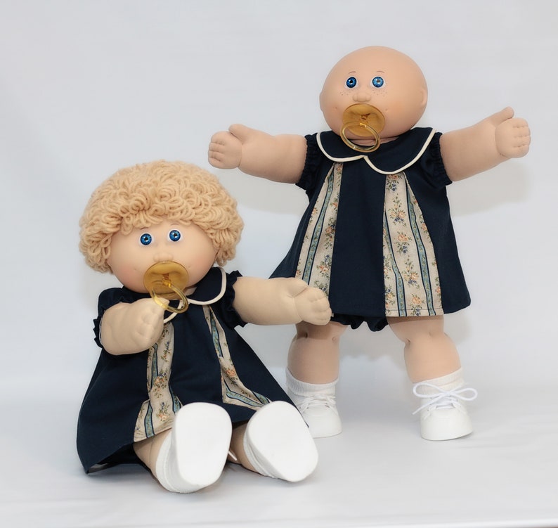 May include: Two Cabbage Patch Kids dolls. One doll is seated, the other is standing. Both dolls have blue eyes, pacifiers, and wear navy blue dresses with floral accents. The seated doll has blonde curly hair, while the standing doll is bald. Both dolls wear white shoes.