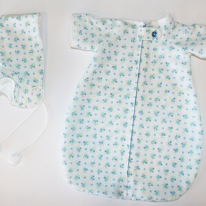 May include: A white baby sleep sack and bonnet set. The fabric is patterned with small blue and green floral designs. The sleep sack has short sleeves and a single blue button closure. The bonnet has ties with white pom-poms.