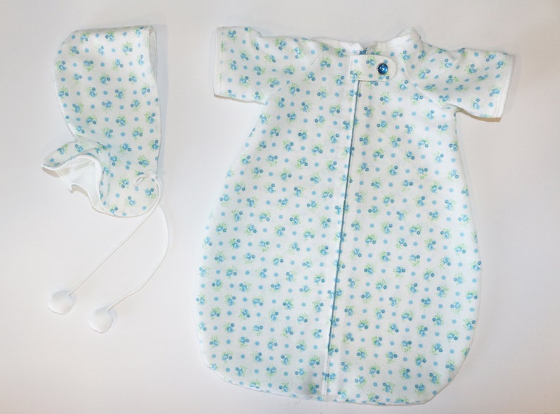 May include: A white baby sleep sack and bonnet set. The fabric is patterned with small blue and green floral designs. The sleep sack has short sleeves and a single blue button closure. The bonnet has ties with white pom-poms.