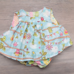 Pode incluir: Um conjunto de vestido e cueca para beb&eacute;. O vestido &eacute; azul claro com um padr&atilde;o de corujas, ramos, flores e nuvens. A cueca tem um padr&atilde;o a condizer. O vestido tem uma saia com folhos.
