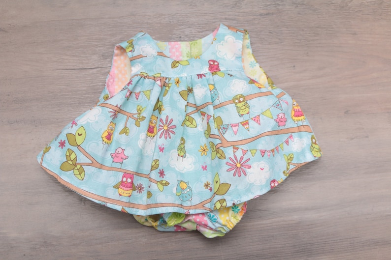 Pode incluir: Um conjunto de vestido e cueca para beb&eacute;. O vestido &eacute; azul claro com um padr&atilde;o de corujas, ramos, flores e nuvens. A cueca tem um padr&atilde;o a condizer. O vestido tem uma saia com folhos.