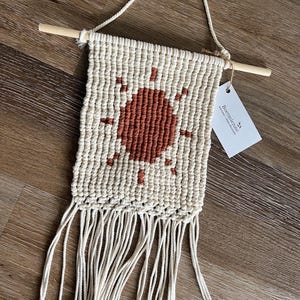 Könnte beinhalten: Ein cremefarbener Makramee-Wandbehang mit einem zentralen, terrakottafarbenen Sonnendesign. Das Stück ist an einem hellen Holzstab mit einer passenden Kordel befestigt und enthält ein weißes Etikett mit der Aufschrift Boetmienne macramé objets décoratifs.
