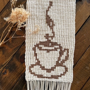 以下が含まれることがあります： ベージュの糸で編まれたマクラメの壁掛けで、茶色の幾何学模様が施されています。上部に木製の棒、下部にフリンジが付いています。