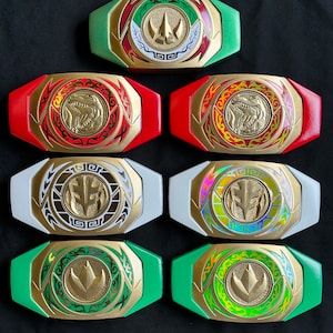 May include: Collection of Power Ranger belt buckles in various colors, including green, red, white, and gold. Each buckle features a central emblem and intricate detailing, with a gold frame and colored accents. The buckles are arranged on a dark surface.