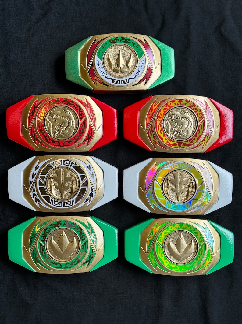 May include: Collection of Power Ranger belt buckles in various colors, including green, red, white, and gold. Each buckle features a central emblem and intricate detailing, with a gold frame and colored accents. The buckles are arranged on a dark surface.