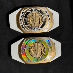 May include: Two white and gold Power Ranger belt buckles. The center emblem is gold with a white background. One buckle has a holographic center. The buckles are on a black surface.