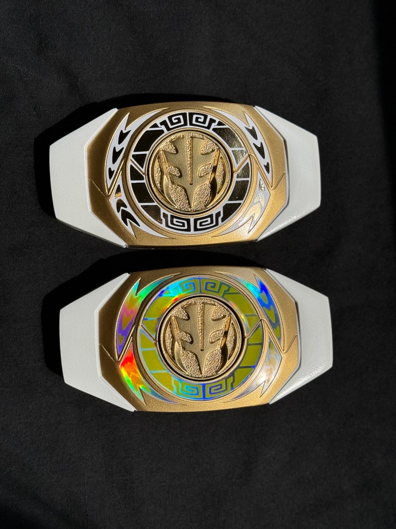 May include: Two white and gold Power Ranger belt buckles. The center emblem is gold with a white background. One buckle has a holographic center. The buckles are on a black surface.