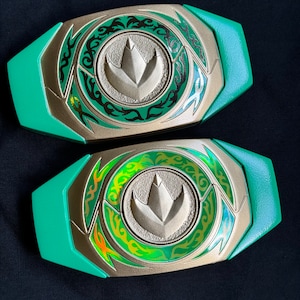 Puede incluir: Dos hebillas de cinturón Power Ranger verdes y doradas. Cada hebilla tiene un emblema central con una huella de dinosaurio estilizada, rodeada por un diseño decorativo verde y dorado. Las hebillas están sobre un fondo oscuro.