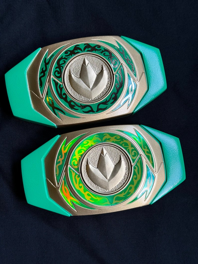 May include: Two green and gold Power Ranger belt buckles. Each buckle has a central emblem with a stylized dinosaur footprint, surrounded by a decorative green and gold design. The buckles are set against a dark background.