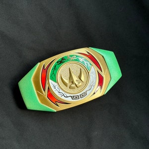 May include: A green and gold Power Rangers belt buckle with a central emblem featuring a gold horn-like design. The buckle has red, white, and green accents and is set against a black background.