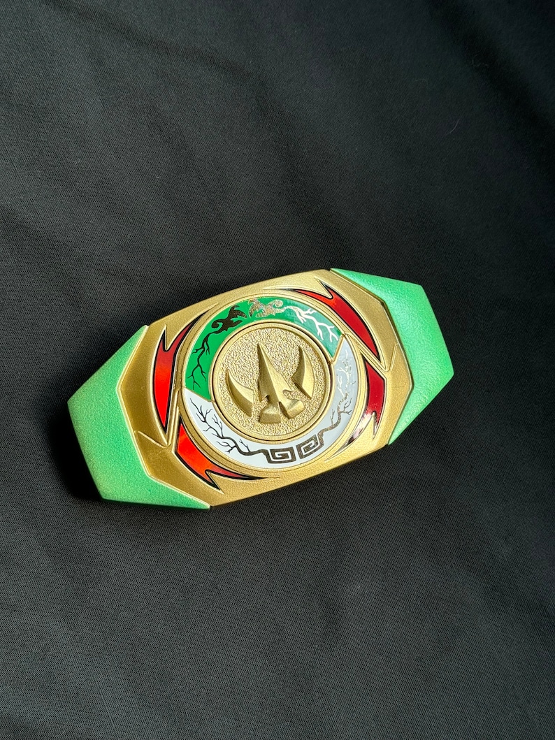 May include: A green and gold Power Rangers belt buckle with a central emblem featuring a gold horn-like design. The buckle has red, white, and green accents and is set against a black background.