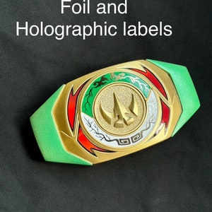 May include: A gold, green, red, and white decorative belt buckle with the text "Drakkon has both Foil and Holographic labels". The buckle has a central emblem with a three-pronged design and is surrounded by intricate patterns.