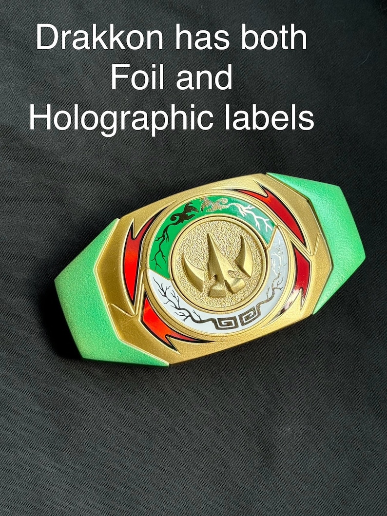May include: A gold, green, red, and white decorative belt buckle with the text "Drakkon has both Foil and Holographic labels". The buckle has a central emblem with a three-pronged design and is surrounded by intricate patterns.