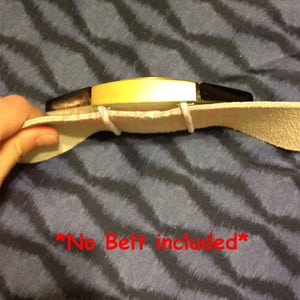May include: A gold-colored belt buckle with black accents is attached to a light tan leather belt. The text "*No Belt included*" is written in red. The buckle is held together with white elastic.