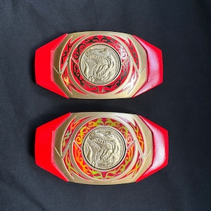 May include: Two red and gold Power Ranger belt buckles. Each buckle features a gold center with a dinosaur emblem, surrounded by a gold and red design. The buckles are set against a black background.