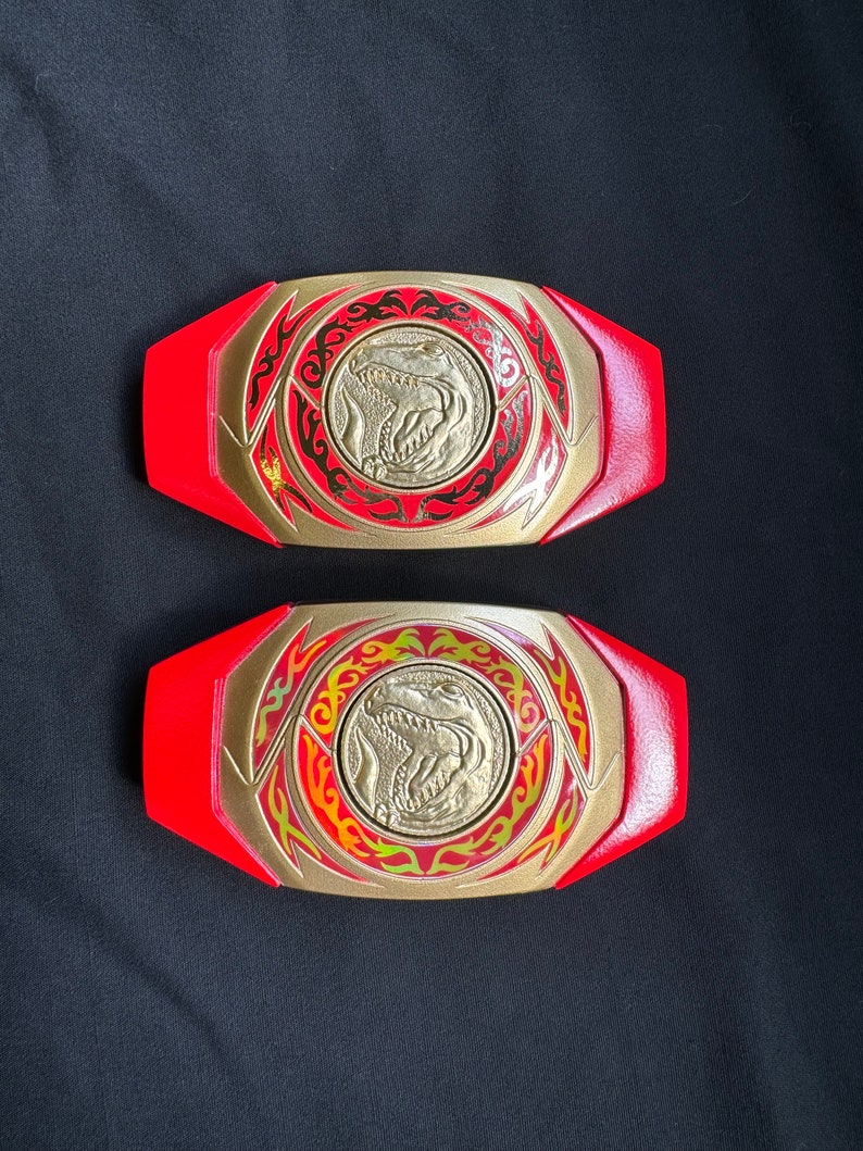 May include: Two red and gold Power Ranger belt buckles. Each buckle features a gold center with a dinosaur emblem, surrounded by a gold and red design. The buckles are set against a black background.