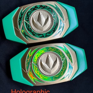 May include: Two prop belt buckles with a gold and green design. One has gold foil accents, and the other has holographic accents. The buckles have a central emblem and are labeled "Example", "Gold Foil", and "Holographic".