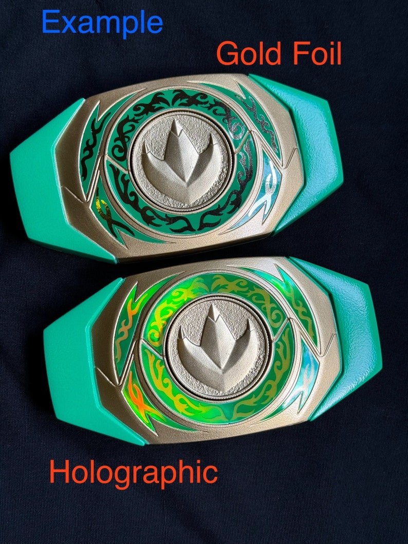 May include: Two prop belt buckles with a gold and green design. One has gold foil accents, and the other has holographic accents. The buckles have a central emblem and are labeled "Example", "Gold Foil", and "Holographic".
