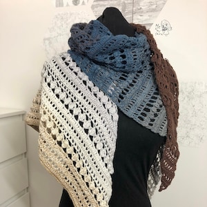 Puede incluir: Un chal de ganchillo en tonos marrones, beige y azules. El chal está hecho con un delicado patrón de encaje y tiene un borde festoneado.