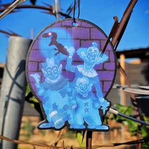 May include: A round car air freshener with a purple and blue background. Three blue cartoon clowns are depicted on the air freshener. The clowns are wearing colorful costumes and have exaggerated features. The air freshener has a string for hanging.
