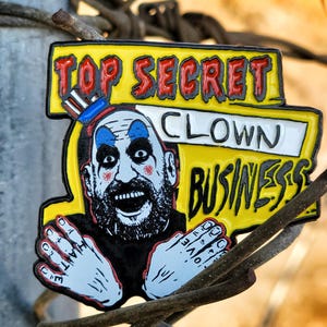 Puede incluir: Un pin de esmalte amarillo con una cara de payaso de dibujos animados y el texto "Top Secret Clown Business". El payaso lleva un sombrero a rayas azul y blanco y tiene una nariz roja.