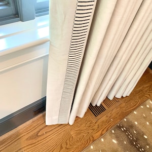 May include: Cream-colored curtain panels with a black striped border. The curtains are hanging near a window with a white sill and a dark gray frame. The floor is hardwood with a deer-patterned rug.