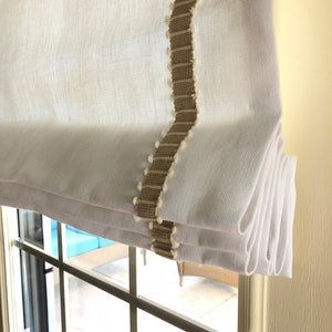 May include: White roman shade with a brown and white trim. The shade is pulled up and folded in a series of pleats.
