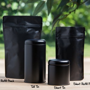 K&ouml;nnte beinhalten: Vier schwarze Beh&auml;lter zum Aufbewahren von Tee oder anderen losen Blattwaren. Zwei hohe Standbeutel und zwei runde Dosen. Die Beutel sind mit "Tall Refill Pouch" und "Short Refill Pouch" beschriftet. Die Dosen sind mit "Tall Tin" und "Short Tin" beschriftet.