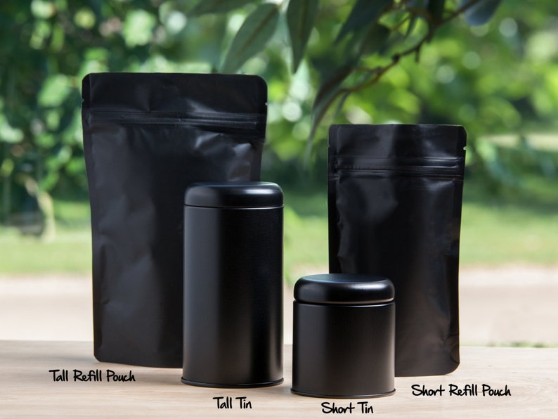 K&ouml;nnte beinhalten: Vier schwarze Beh&auml;lter zum Aufbewahren von Tee oder anderen losen Blattwaren. Zwei hohe Standbeutel und zwei runde Dosen. Die Beutel sind mit "Tall Refill Pouch" und "Short Refill Pouch" beschriftet. Die Dosen sind mit "Tall Tin" und "Short Tin" beschriftet.