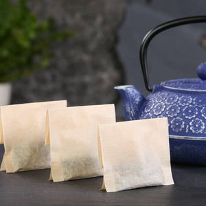 May include: Three tea bags sit in front of a blue teapot with a black handle. The teapot has a floral pattern. The tea bags are light brown and rectangular. A green plant in a white pot is in the background.