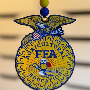 May include: A yellow and blue FFA emblem with an eagle, owl, and the words "AGRICULTURAL EDUCATION" and "FFA". The emblem is hanging from a black string with yellow and blue beads.
