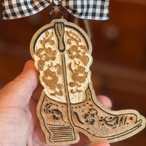 May include: A decorative ornament shaped like a cowboy boot, featuring intricate floral designs and a glittery finish. The boot is outlined in black and hangs from a twine loop with a black and white checkered bow.