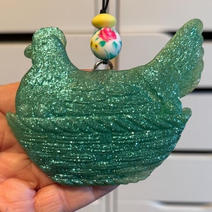 May include: A teal glittery chicken-shaped ornament. The ornament is held by a black cord with a floral bead and a yellow bead. The chicken has a detailed wing and tail feathers. The ornament is held in a hand.