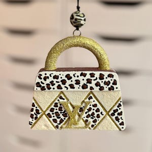 May include: A decorative handbag ornament with a gold glitter handle and a cream-colored body. The handbag features a leopard print design and a gold logo. The ornament is suspended by a black string and a zebra-striped bead.