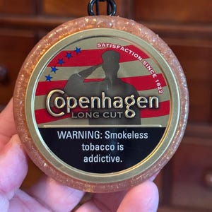 May include: A round, amber-colored Copenhagen Long Cut tobacco container with a black cord loop. The label features a saluting silhouette against a red and blue American flag design. Text includes "Satisfaction Since 1822" and a warning about addictive smokeless tobacco.