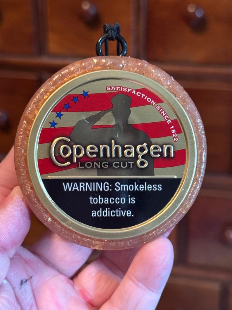 May include: A round, amber-colored Copenhagen Long Cut tobacco container with a black cord loop. The label features a saluting silhouette against a red and blue American flag design. Text includes "Satisfaction Since 1822" and a warning about addictive smokeless tobacco.