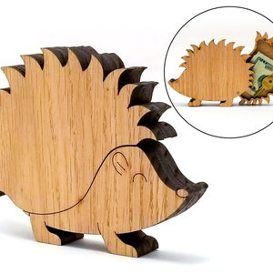 Peut inclure: Une boîte en bois en forme de hérisson avec un visage souriant et un dos épineux. La boîte est en bois brun clair avec des accents brun foncé. Un encart circulaire montre la boîte ouverte, révélant un compartiment caché avec de l'argent et une clé.