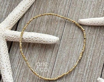 以下が含まれることがあります： 木製の表面に「arie & co」の文字が入った繊細なゴールドビーズのブレスレット。白いヒトデに囲まれ、ビーチのような美しさ。ブレスレットの周囲は約18cmです。