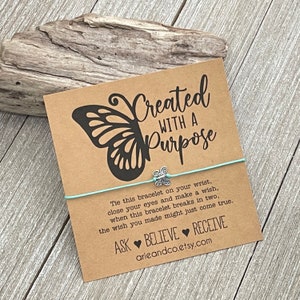 May include: A light blue string bracelet with a silver butterfly charm. The bracelet is tied around a card with a butterfly graphic and the text "Created with a Purpose". The card also includes the text "Tie this bracelet on your wrist, close your eyes and make a wish, when this bracelet breaks in two, the wish you made might just come true. Ask Believe Receive arieandco.etsy.com"