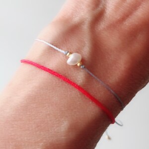 Puede incluir: Dos pulseras en una mu&ntilde;eca. Una es de hilo rojo y la otra es de hilo gris con una perla blanca.