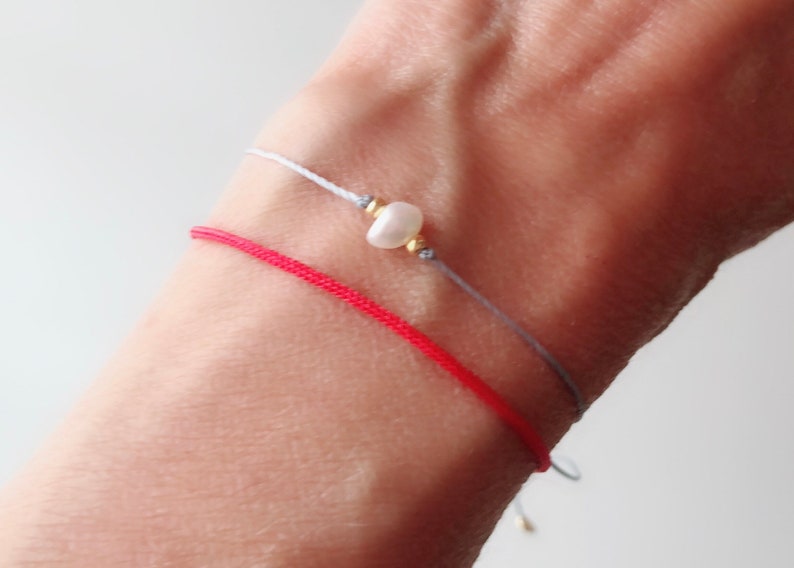 Puede incluir: Dos pulseras en una mu&ntilde;eca. Una es de hilo rojo y la otra es de hilo gris con una perla blanca.