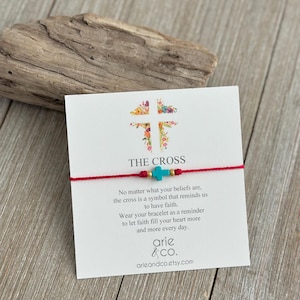 May include: A red string bracelet with a turquoise cross charm and gold beads. The bracelet is displayed on a white card with the text "THE CROSS" and a floral cross design. The card also includes text about faith.