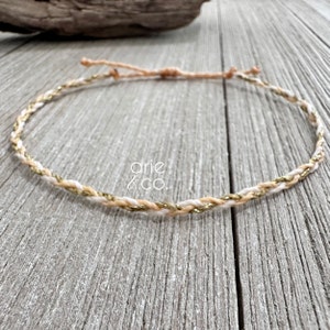 May include: A braided cord necklace with gold and cream colored threads. The necklace is on a wooden surface.