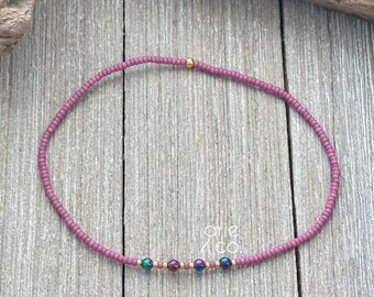 May include: A delicate pink beaded bracelet with iridescent accents and gold beads. The bracelet is on a wooden surface.