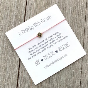 May include: A white card with pink string tied to it. The string has a small gold rose charm. The card reads "A Birthday Wish for you" and includes the text "Tie this bracelet on your wrist, close your eyes and make a wish, when this bracelet breaks in two, the wish you made might just come true. ASK BELIEVE RECEIVE arieandco.etsy.com"