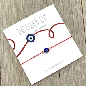 May include: Red string anklet with a blue evil eye charm. The anklet is on a white card with the text "The Lucky Eye anklet bracelet" and "arieandco.etsy.com".