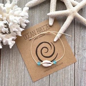 May include: A white seashell charm with two light blue glass beads on a beige string bracelet. The bracelet is tied with a knot and has a tag that reads "Ocean Breeze" and "Anklet bracelet".